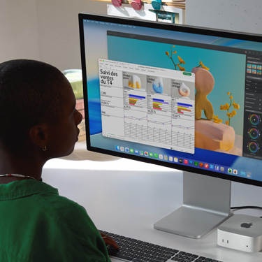 Sur iMac et Mac mini, une personne assise travaille sur une feuille de calcul Excel portant sur le suivi des ventes du quatrième trimestre. Derrière la feuille de calcul Excel se trouve une fenêtre de Pixelmator Pro et l’arrière-plan de macOS Tahoe.
