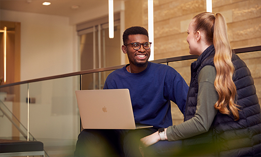 Chidi zit met een MacBook op schoot en praat met een collega.