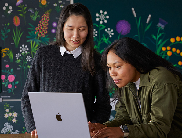 Two Apple Marketing team members working together on a MacBook in front of posters designed by their team.