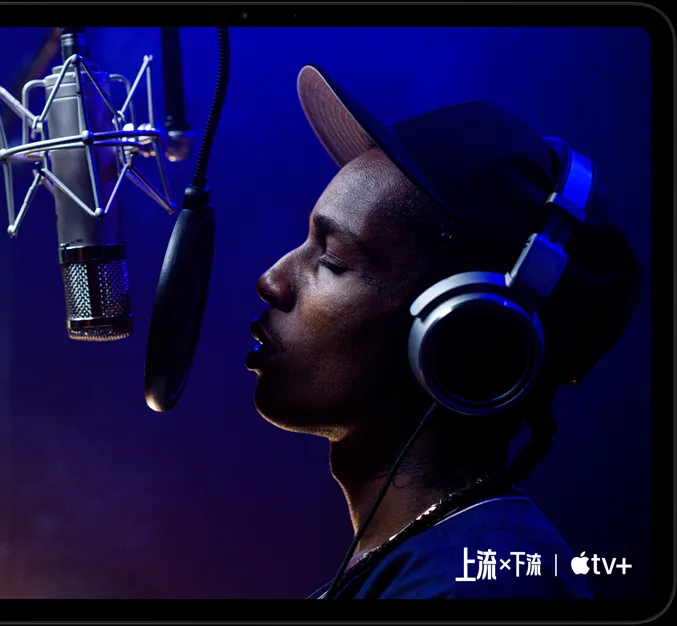 橫向放置的太空黑色 iPad Pro 機身正面，展示圓角設計與黑色顯示器邊框，顯示器以 Apple TV+ 播放 Apple TV+ 原創電影上流 × 下流中的一幕，由 ASAP Rocky 飾演的 Yung Felon 正在對著麥克風唱饒舌歌