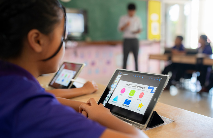 Close-up of a student at their desk, using an iPad. On screen, there are colorful shapes displayed with the title, "Meet the shapes."