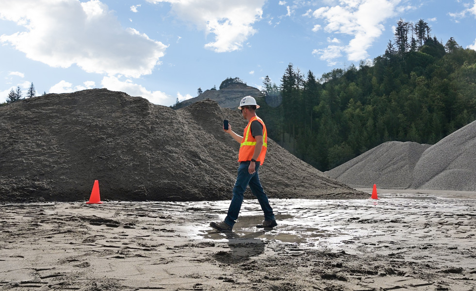 Un homme de terrain évalue un chantier, un iPhone à la main, en marchant sur un sol boueux