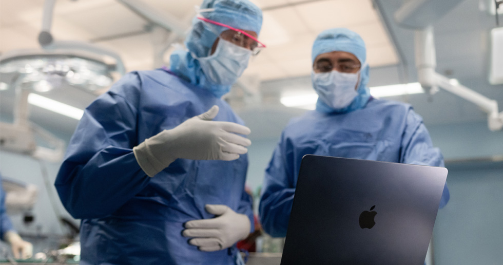Deux chirurgiens dans une salle d’opération consultent un MacBook, l’un d’eux fait un geste de la main tandis que l’autre observe