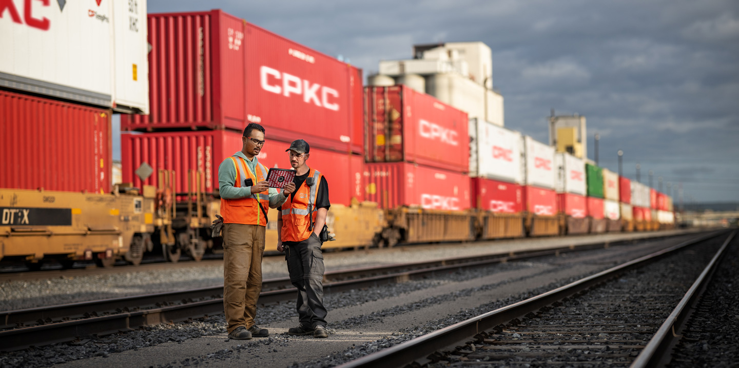 Deux cheminots consultent un iPad dans une gare de triage à côté d’un train immobilisé chargé de conteneurs