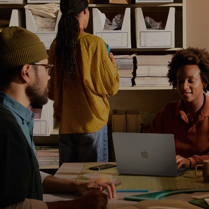 Trois personnes dans un bureau, deux personnes assises semblent faire allusion au contenu affiché sur un écran de MacBook tandis qu’une troisième personne fait face à un tableau noir sur lequel figurent plusieurs graphiques