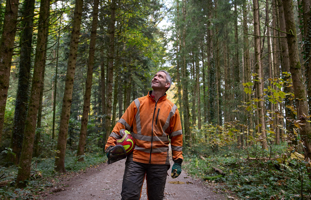 Man in een oranje veiligheidsvest met een veiligheidshelm op staat op een bospad in een duurzaam beheerd bos in Zwitserland